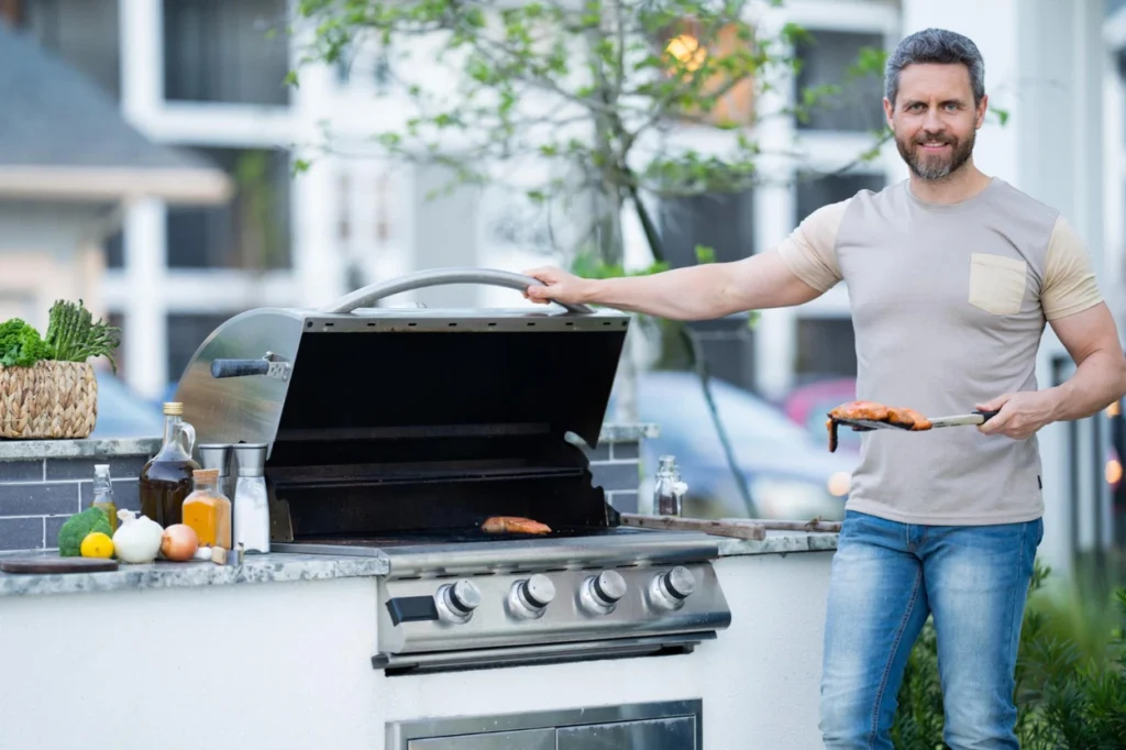 Homem sorrindo ao lado de sua churrasqueira a gás de embutir em uma bancada de granito, demonstrando a praticidade de um espaço gourmet.
